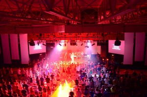 A large crowd watches a live concert indoors with red lighting and video screens displaying lyrics.