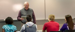 A teacher stands at the front of a classroom holding a tablet, smiling, and speaking to four students seated at desks. The students, who are pursuing their KCU Degrees, listen attentively.