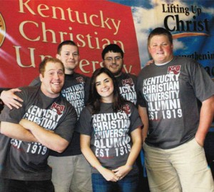 Five young adults wearing "Kentucky Christian University Alumni" t-shirts pose together for a group photo in front of a banner with the university's name.