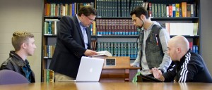 Three men converse with a professor at a podium in a library; a laptop is on the table.
