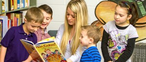 A young woman reads "Yum, Cookies!" to a group of four children in a library setting. The children attentively gather around, engaging with the book.