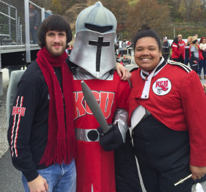 Three people are posing together, one in a knight's costume, likely a mascot, and two others in university-branded attire, suggesting school spirit at a Kansas City University (KCU) event celebrating KCU degrees.