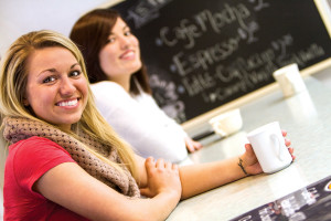 Two women sitting at a coffee shop counter, smiling and holding white coffee mugs, with a chalkboard menu in the background, chat about their upcoming admissions interviews.
