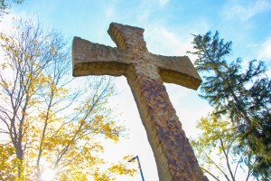 A large, rugged cross statue stands outdoors, surrounded by trees with yellow leaves and a partly cloudy sky.