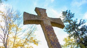 A large stone cross stands against a bright blue sky, surrounded by trees with autumn leaves.