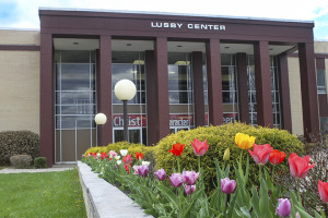 Exterior view of a building with large windows, brick columns, and a row of colorful tulips in the foreground. Signs behind the windows read "Christ," "Character," "Career." The inviting atmosphere hints at a place where admissions play a key role in shaping futures.