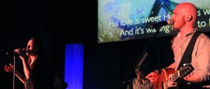 Two musicians perform on stage under blue lighting; a woman sings while a man plays an acoustic guitar and sings. A projected screen in the background, reminiscent of instructional presentations for KCU Degrees, displays partial lyrics.