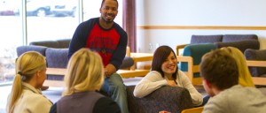 A group of six people, seated in a casual lounge area, engage in a discussion; one individual stands while others are seated in chairs, smiling and talking.