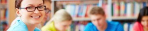 A person wearing glasses and a blue shirt smiles at the camera in a library setting, possibly after a successful admissions interview; several other people are seen in the background, blurred, focusing on their tasks.