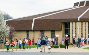 A group of people walk outside a modern church building with a cross, surrounded by greenery and trees.