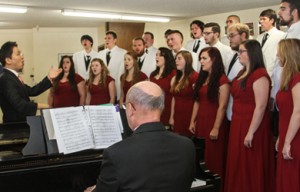 A choir of men in white suits and women in red dresses passionately sing as a conductor leads them and a pianist plays the piano, reminiscent of a symphony celebrating KCU Degrees.