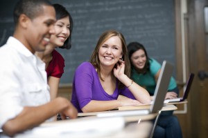 A group of people smiling and conversing around a laptop in a classroom setting with a chalkboard in the background.