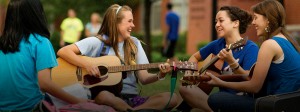 A group of young women sit outdoors, playing guitars and laughing together. They appear to be enjoying a casual, sunny day.