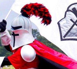 A person wearing a knight costume with a cross on the helmet and red plume is holding a flag adorned with a cross and shield emblem.