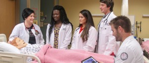 A group of medical trainees stand around a hospital bed with a medical mannequin. One person is seated and using a tablet. All are wearing white lab coats.
