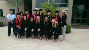 A group of people in graduation gowns and caps pose for a photo outside a building, with two adults standing on the sides.