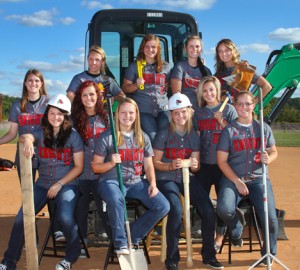 A group of young women in matching jerseys labeled "Knights" pose on and around construction equipment, holding various tools including a shovel, hammer, and axe, on a sunny day at a baseball field.