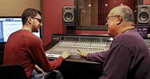 Two men are working at an audio mixing console in a recording studio, with a computer screen displaying audio tracks in the background. Their expertise is evident, thanks to their KCU Degrees in sound engineering.
