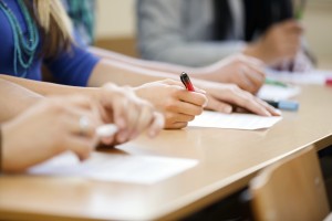 Close-up of students sitting at desks, writing on paper with pens in a classroom setting.