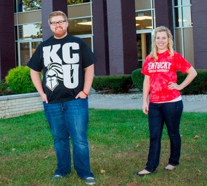Two people stand outdoors on grass in front of a building. The person on the left wears a black KCU t-shirt. The person on the right wears a red Kentucky Christian University t-shirt.
