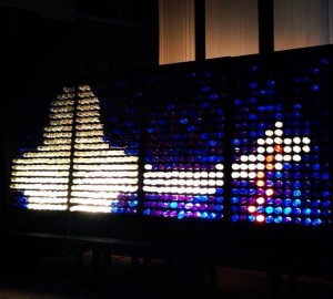 A colorful abstract LED light display featuring a large white triangle on the left and a white cross on the right against a dark background.