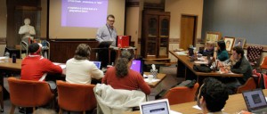 A man stands at a podium and presents a slideshow on KCU Degrees to a room of seated people using laptops and notebooks.