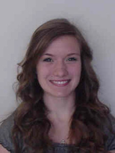 A person with long, wavy brown hair smiles at the camera against a plain background.