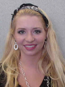 A woman with long blonde hair, wearing a black headband, dangling earrings, and a necklace, smiles at the camera.