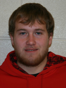 A close-up photo of a person with short, light brown hair, a short beard, and blue eyes, wearing a red and black plaid shirt. A light-colored brick wall serves as the background.