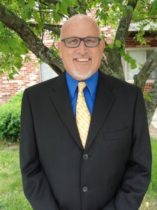 A Kentucky Christian University Alumni, wearing a dark suit with a blue shirt and a yellow patterned tie, stands outside near a tree.