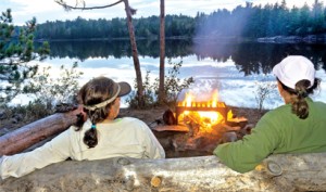 Two people sit on a log by a campfire, facing a lake surrounded by trees at dusk.