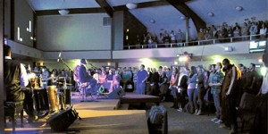 A large group of people stands and watches a music performance in a church-like venue with high ceilings and a second-floor balcony.