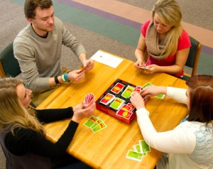 Four people sitting around a table, engaged in a card game with red and green cards. The game box is on the table, and cards are scattered around.