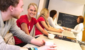 A group of young people sit at a counter with drinks, engaging in conversation. A woman stands behind the counter near a menu board.