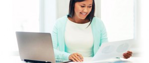 A woman sits at a desk with a laptop, smiling as she looks at a document with charts.