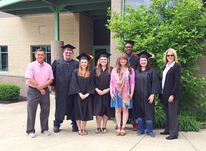 Seven graduates in caps and gowns stand with two adults outdoors near a building with bushes in the background.