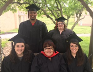 Group of five graduates in caps and gowns standing outdoors, with two standing at the back and three sitting in the front. They are smiling, and a tree and grass are visible in the background.