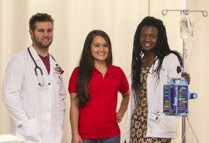 Three medical professionals stand together; two are in white coats with stethoscopes, and the third is in a red shirt. One holds an IV stand with medical equipment.