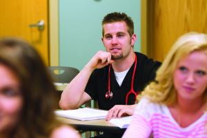 A man with a stethoscope around his neck sits in a classroom, attentively listening and taking notes. Two other people are partially visible in the foreground.