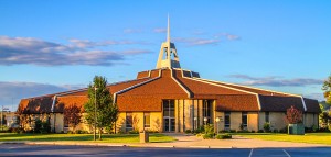 A modern church with a multi-faceted roof and a tall steeple, surrounded by a well-maintained lawn, seen on a sunny day with a clear sky.