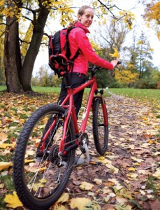 A person in a red jacket with a backpack is standing with a red bike on a leaf-covered path in a park during autumn.
