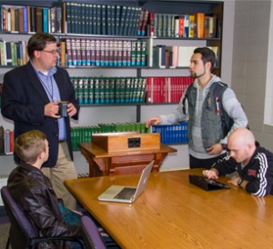 Group of three students and a professor in a library. One student is seated with a laptop, another with a tablet, and the third is standing and talking to the professor who holds a coffee mug.