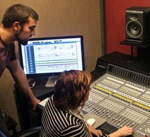 Two people working in a recording studio; one person is standing and looking at a computer screen displaying audio tracks, while the other is seated and operating a large mixing console.