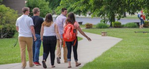 A group of five people, including two women and three men, walk together on a sidewalk through a grassy area with trees in the background. One person wears a red backpack.
