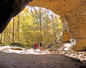 Three people stand under a large rock archway, surrounded by autumnal trees in a forest setting.