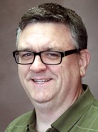 A man with short brown hair and glasses smiling, wearing a green collared shirt, against a plain brown background.