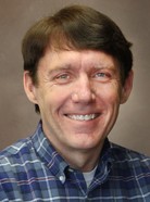 A man with short brown hair, wearing a blue checkered shirt, smiles in a portrait photo with a brown background.