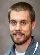 A man with short brown hair, a beard, and a mustache, wearing a gray collared shirt, smiles at the camera.
