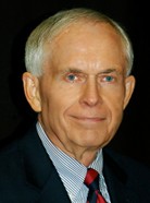 An older man with short gray hair, wearing a dark suit, light blue shirt, and red tie, against a dark background.