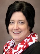 A person with short dark hair, wearing a black top and red and white patterned scarf, smiling at the camera against a neutral brown background.